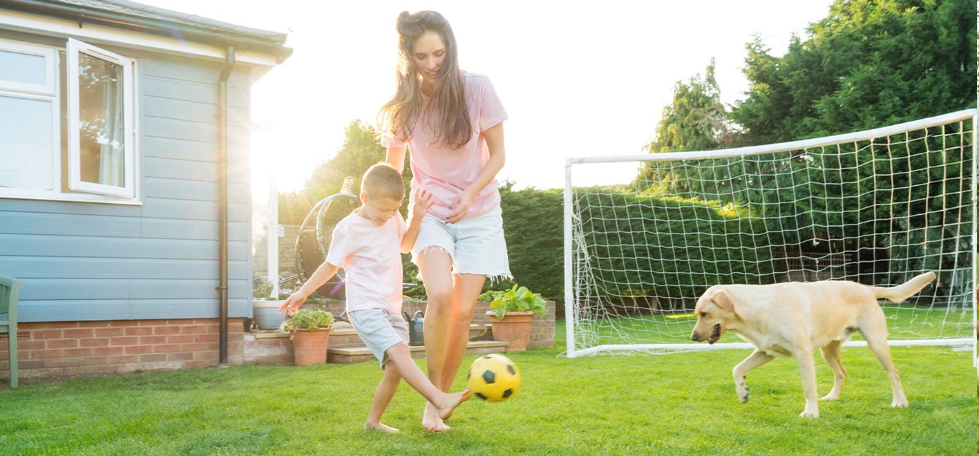 Solar-Maulwurf-Vertreiber SuperCat – Entwickelt für ruhiges, komfortables Leben im Freien. Lifestyle-Bild zeigt eine Frau und ein Kind, die mit einem Fußball auf einer Rasenfläche im Garten spielen, mit einem Hund in der Nähe sowie einem Haus und einem Fußballtor im Hintergrund.
